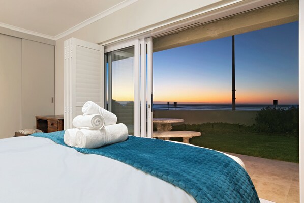 A bedroom with a bed, a blue blanket, and white towels. There is a sliding glass door leading to a balcony with a view of the beach and ocean at sunset. - Melkbos Aan See (Cape Town)