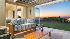 A sunlit patio with a wooden bench, a striped sofa, and a glass door leading to a grassy area with a view of the ocean. - Melkbos Aan See (Cape Town)