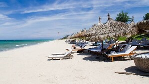 A beach with rows of sun loungers and thatched umbrellas, a clear blue sky, and the ocean in the background. - Hoian Royal Villa (Da Nang)