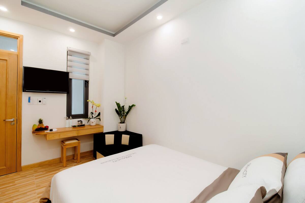 A hotel room with a bed, a desk with a chair, a small table with a plant, and a television mounted on the wall.