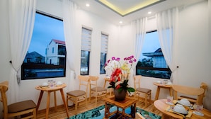 A room with wooden furniture, a table with a vase of flowers, and large windows with curtains. - Hoian Royal Villa (Da Nang)
