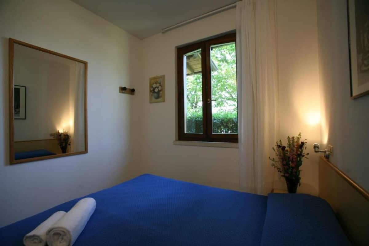 A bedroom with a bed, a window with a view of greenery, a mirror, and a wall-mounted light fixture.