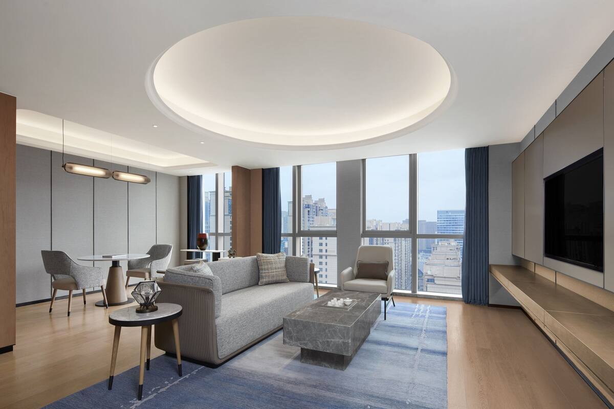 A modern living room with a large sofa, a coffee table, and a TV. There are two chairs and a small table. The room has large windows and a circular ceiling light.