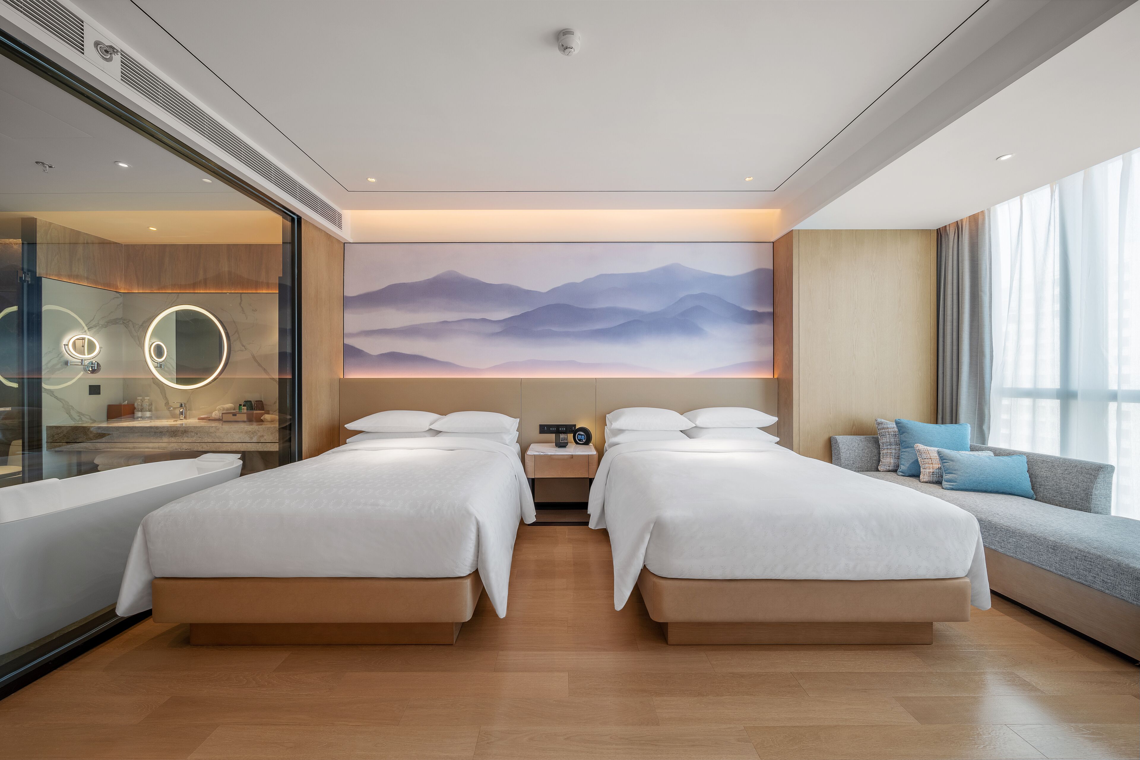 A modern hotel room with two beds, a large window, a wall-mounted artwork, and a bedside table with a lamp.