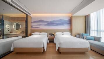 A modern hotel room with two beds, a large window, a wall-mounted artwork, and a bedside table with a lamp.