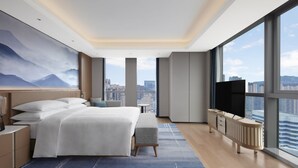 Ein modernes Hotelzimmer mit einem großen Bett, einem Fernseher, Blick auf die Stadt und einem großen Fenster.
