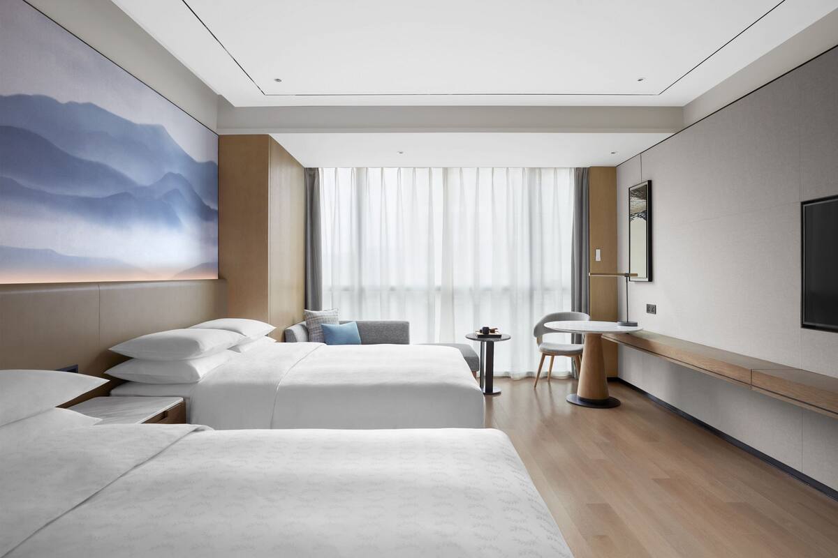 A hotel room with two beds, a desk, a chair, a TV, and a large window with curtains.