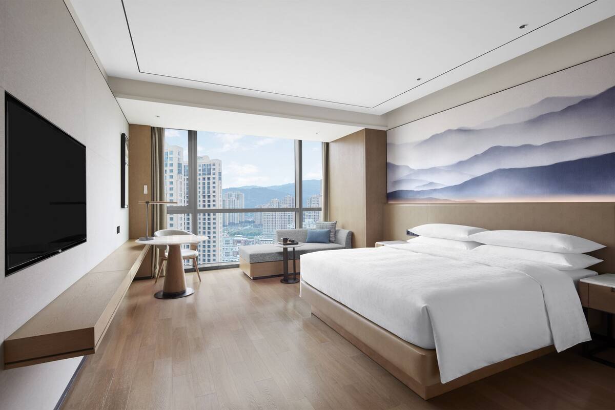 A modern hotel room with a large bed, a flat-screen TV, a seating area with a table, and a city view through large windows.