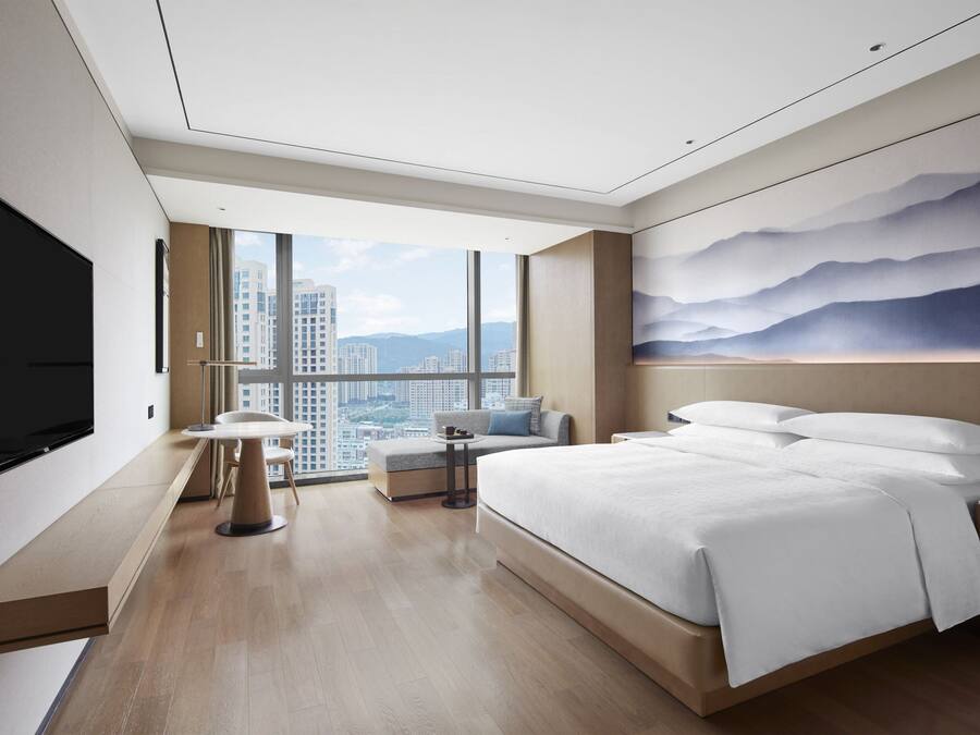 Una habitación de hotel moderna con una cama grande, un televisor de pantalla plana, una zona de estar con una mesa y vistas a la ciudad a través de ventanas amplias.