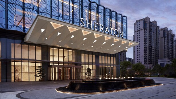 Ein Sheraton Hotel mit moderner Glasfassade, einem markanten Eingang mit geometrischer Markise und einem begrünten Bereich mit Wasserspiel.