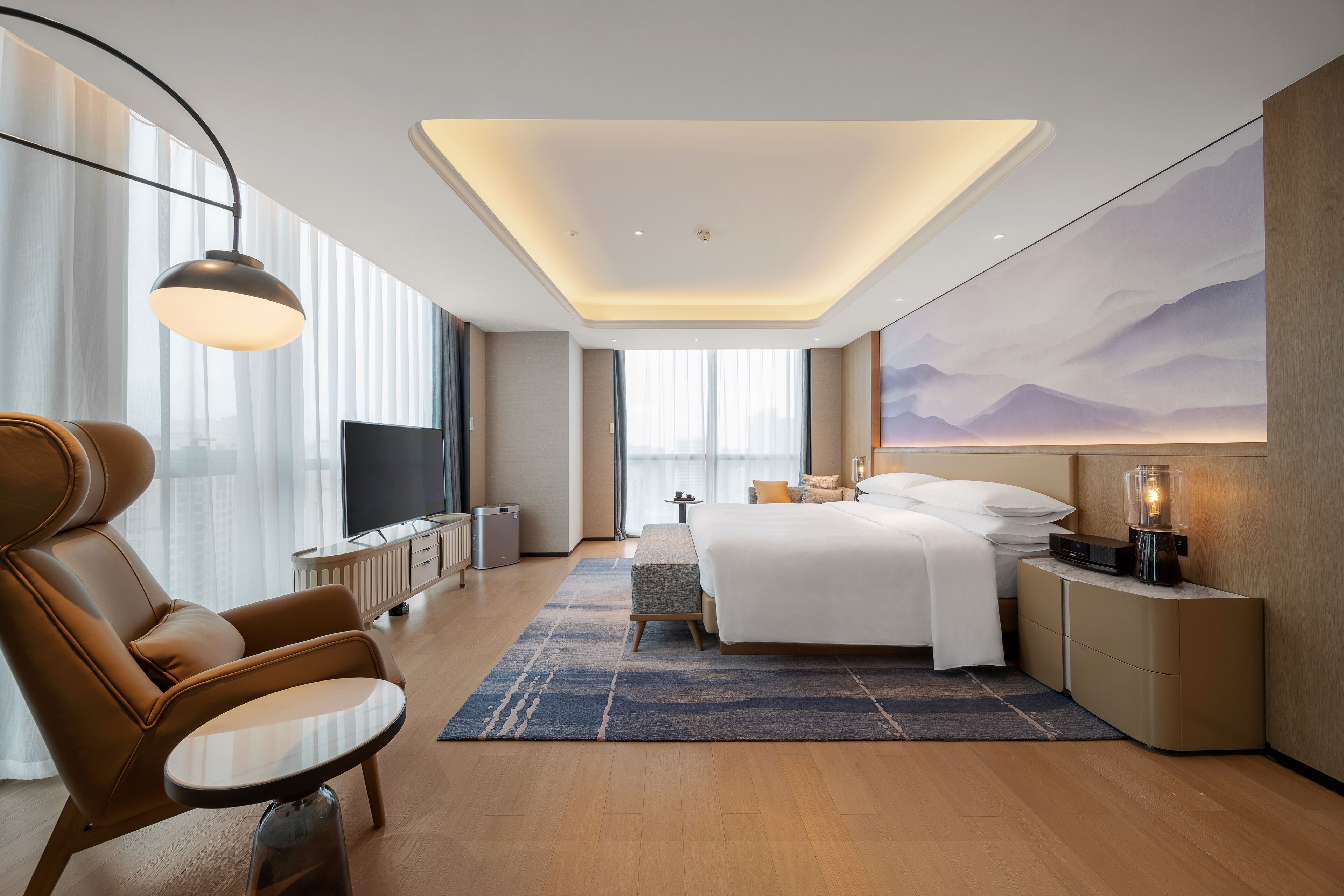 Ein modernes Hotelzimmer mit einem großen Bett, einer Sitzecke inklusive Sessel und Tisch, einem Fernseher und einem großen Fenster mit Vorhängen.