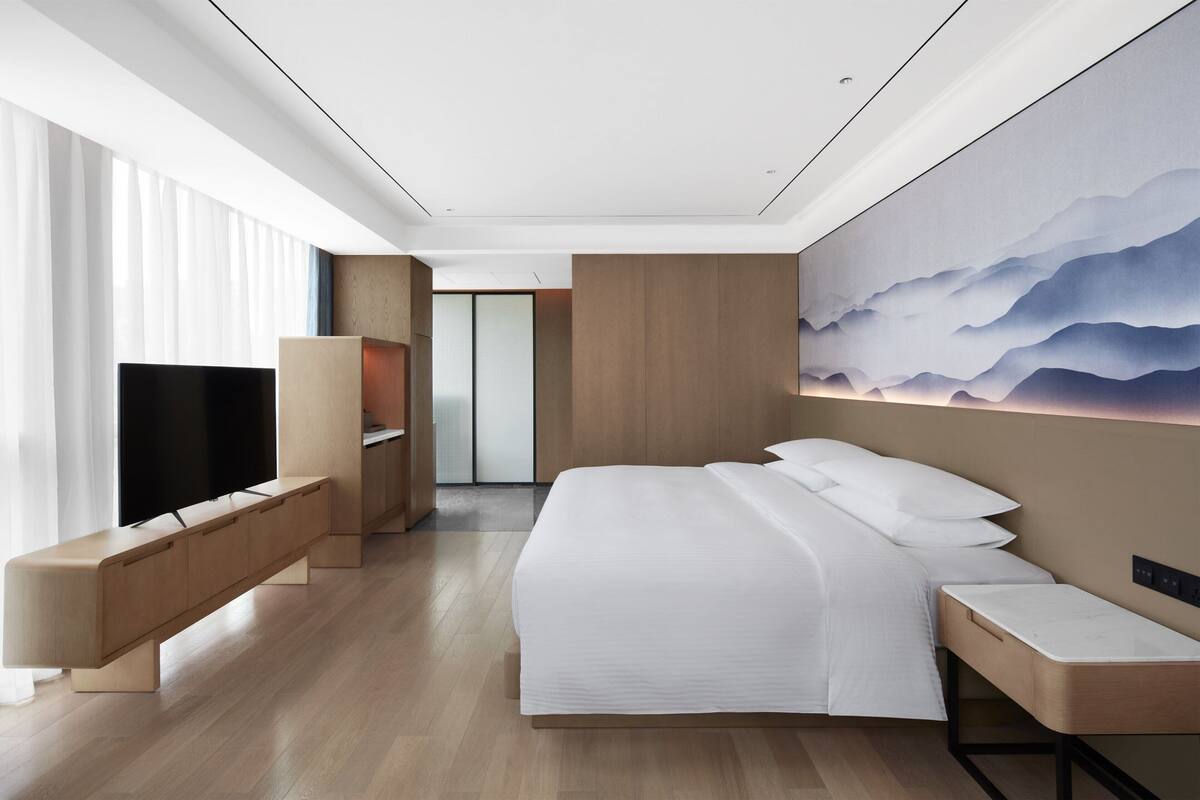 A modern hotel room with a large bed, a TV, a seating area, and a view of mountains through the window.