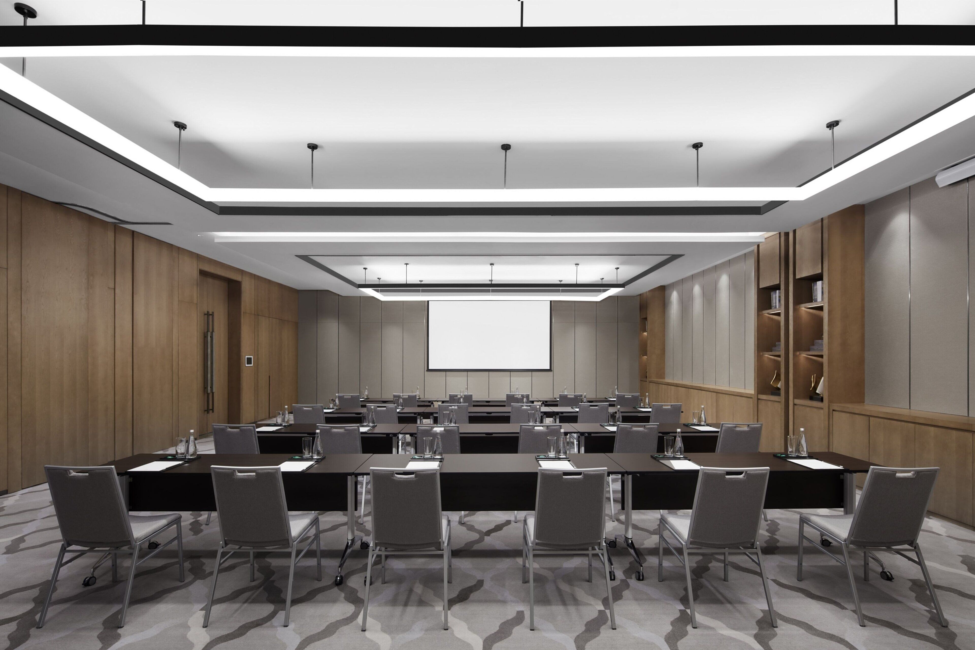 Un salón de conferencias con una larga mesa, sillas, una pantalla de proyector y piso alfombrado.