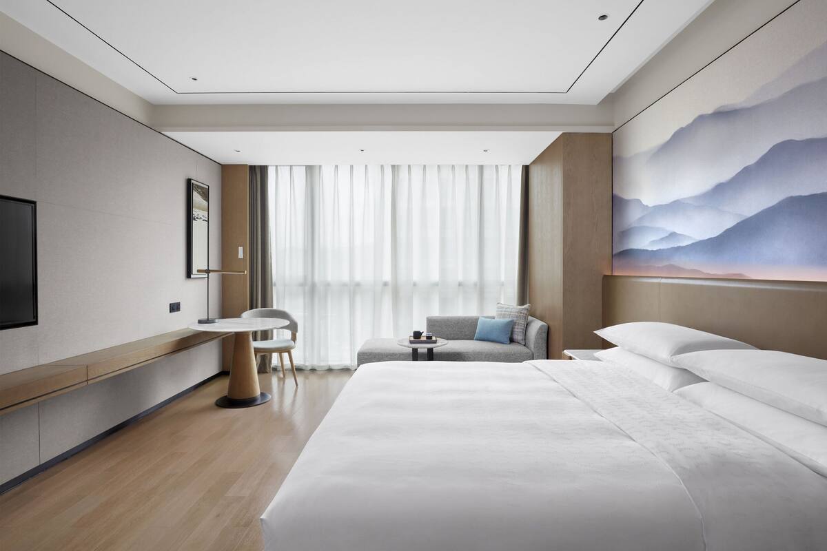 A modern hotel room with a large bed, a desk, a chair, and a sofa.