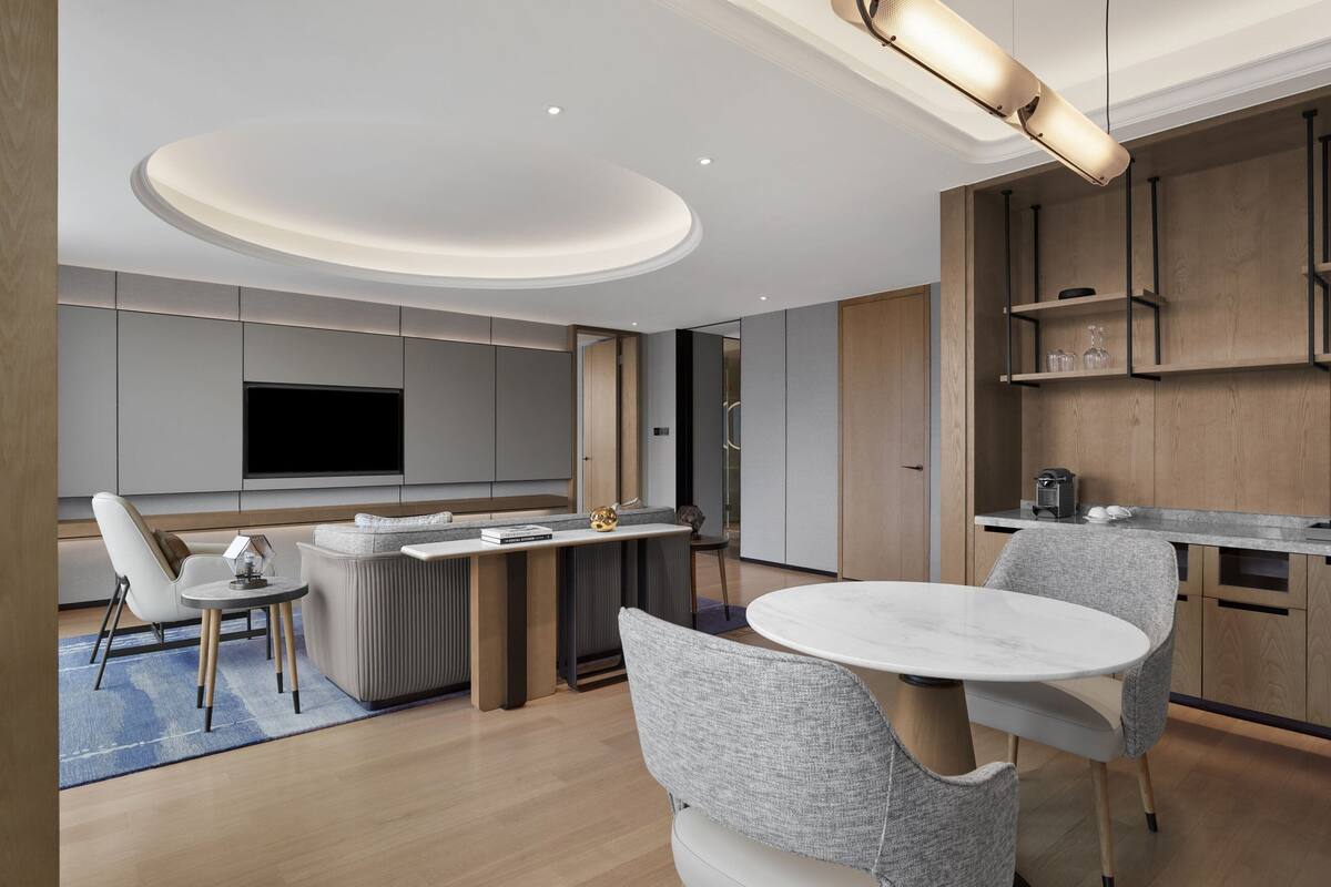 A modern living room with a flat-screen TV, a dining area with a round table, and a kitchen with wooden cabinets.
