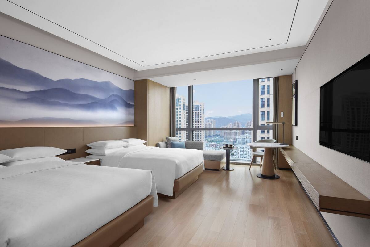 A modern hotel room with two beds, a seating area, and a large window offering a city view.