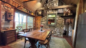A rustic kitchen with a stone wall, wooden beams, and a large dining table. - Enjoy this 100+ year old cabin in the gated Wolf Laurel Resort community. (MARS HILL)