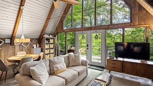 Un ampio soggiorno con soffitto alto, ampie finestre, travi a vista in legno, un divano, una televisione, un tavolo da pranzo e una libreria.
