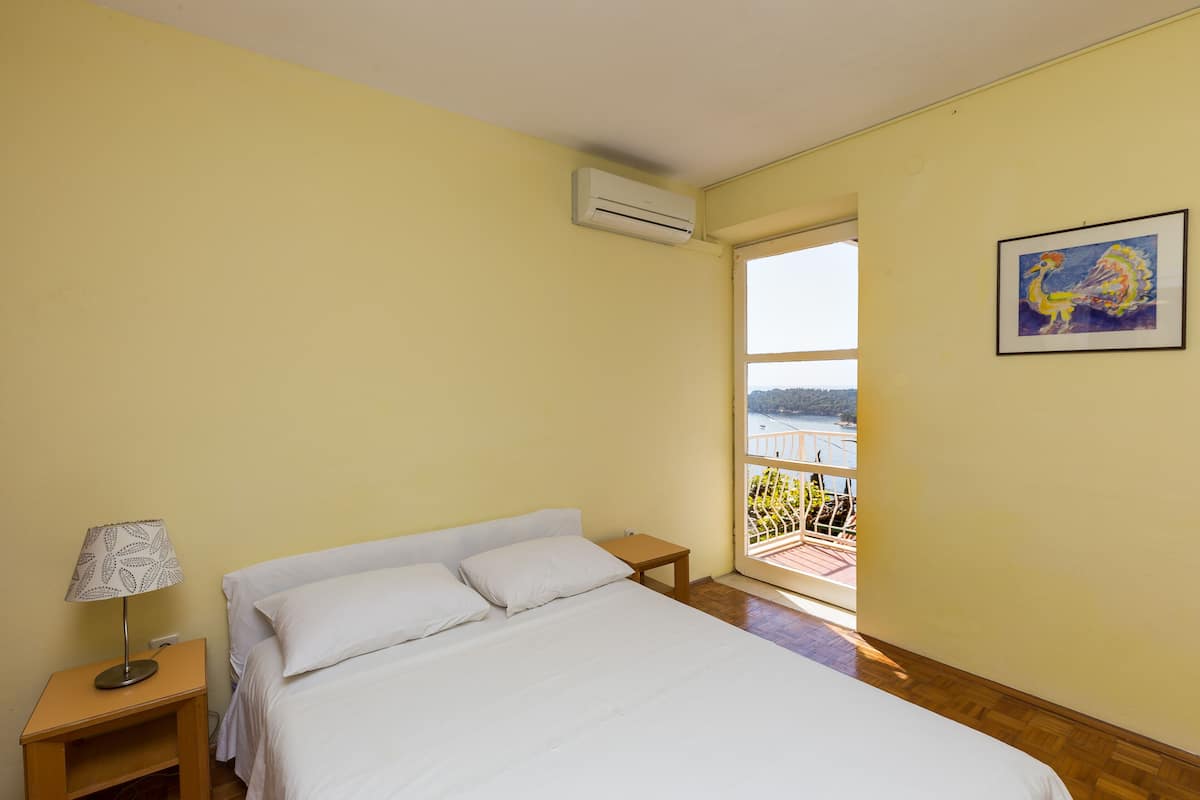 Une chambre à coucher comprenant un lit, une table de chevet, un appareil de climatisation et une fenêtre donnant sur un balcon.