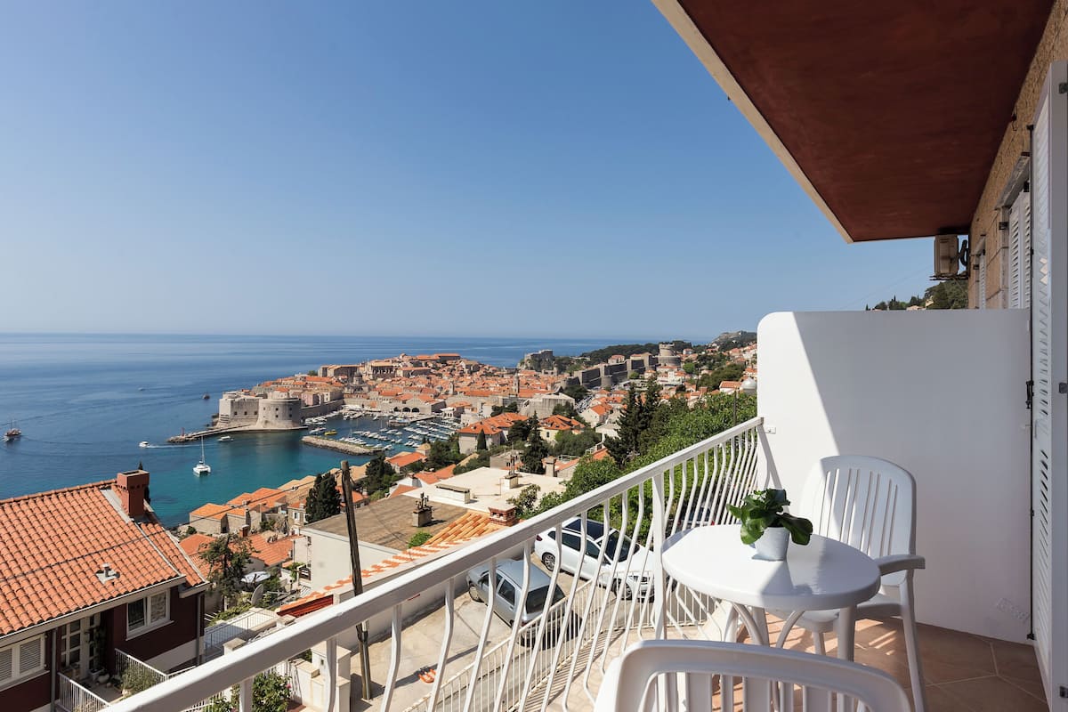 Un balcon avec des chaises blanches, un pot de fleurs et une vue sur une ville côtière, avec ses bâtiments et son port.