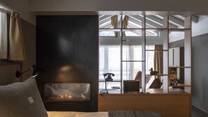 Una habitación de hotel moderna con chimenea, una cama con almohadas, un escritorio y vistas a la ciudad a través de amplios ventanales.