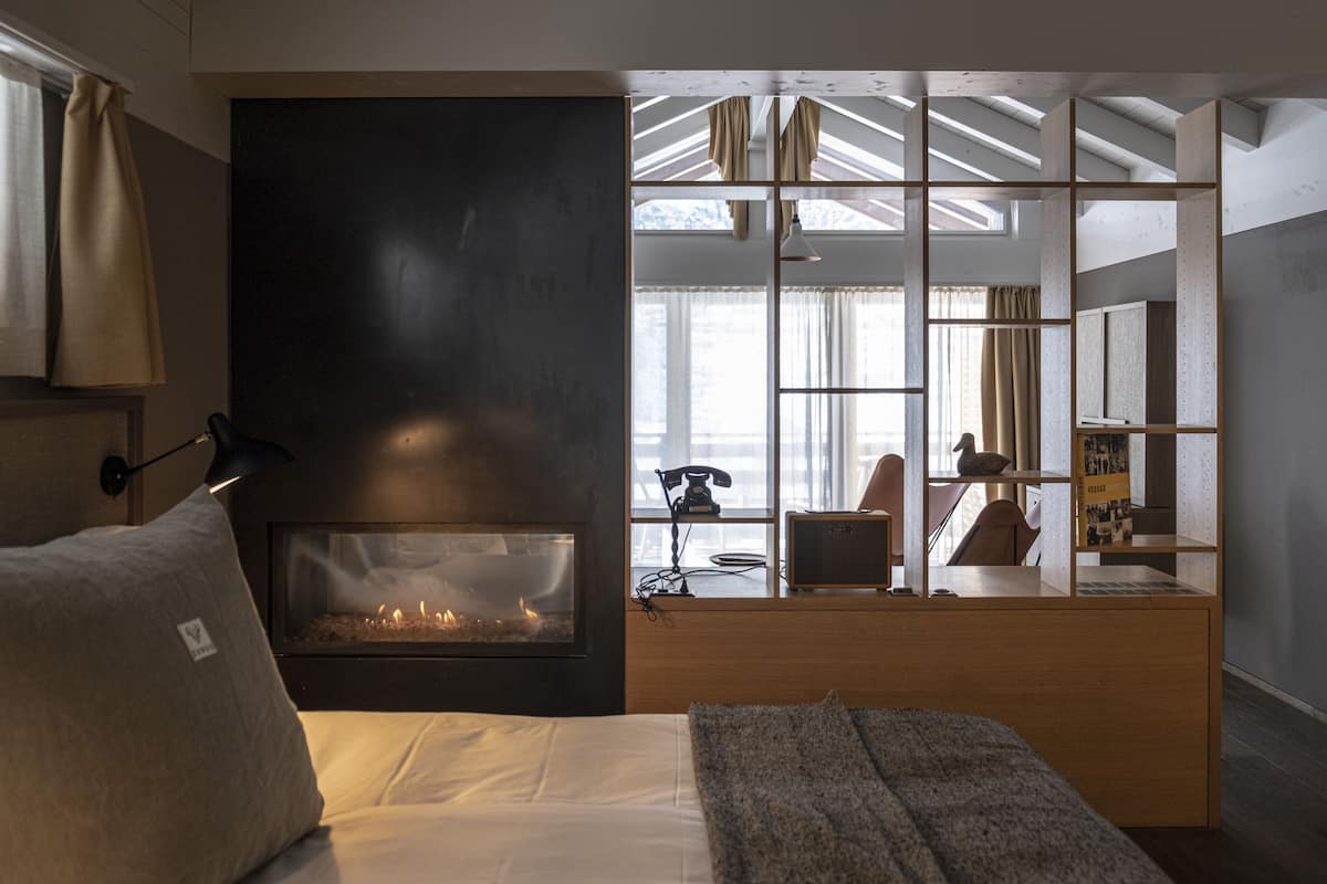 A modern hotel room with a fireplace, a bed with pillows, a desk, and a view of the city through large windows.
