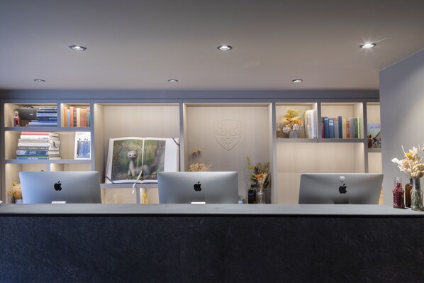 A reception desk with three Apple iMac computers, a bookshelf with books and decorative items, and a wall with a heart-shaped design.