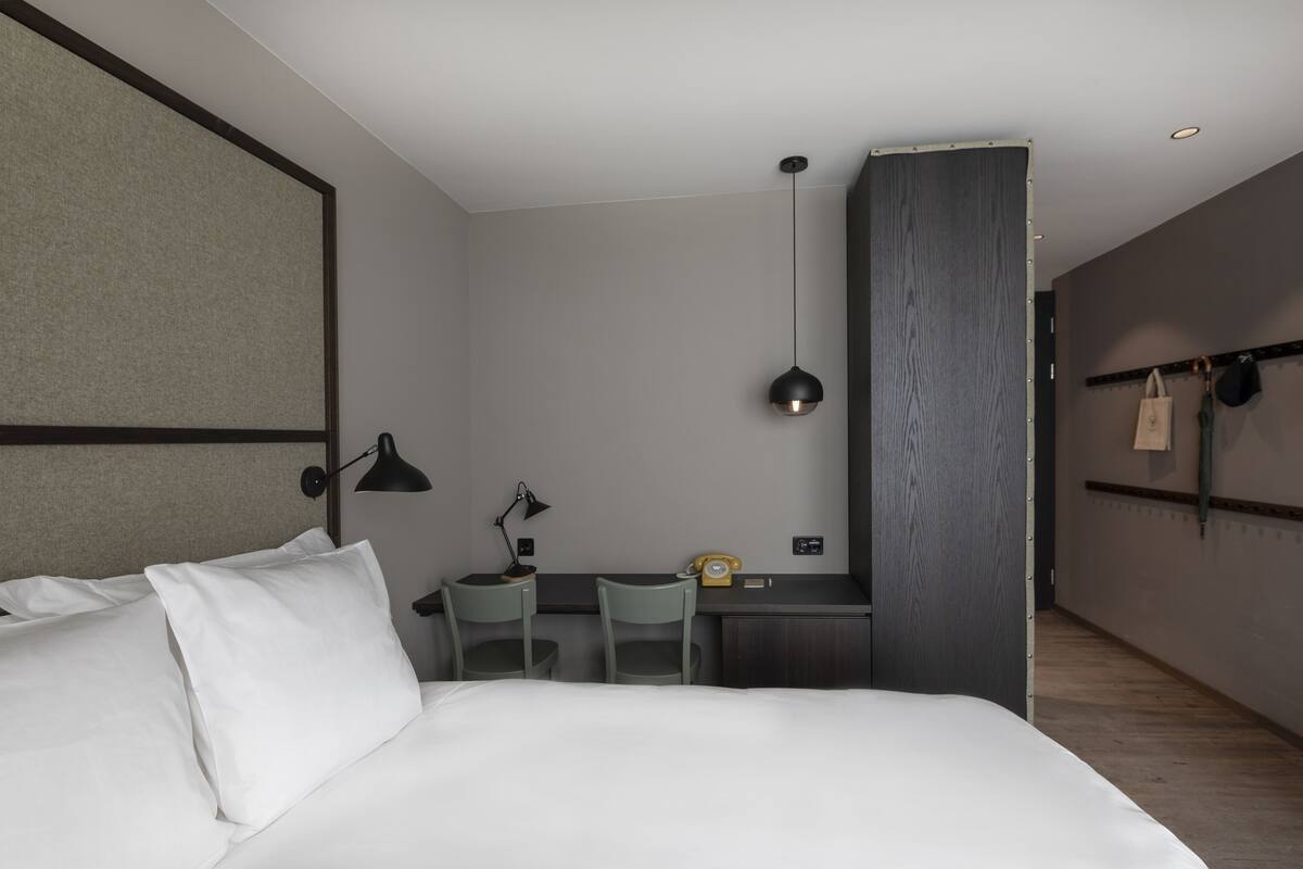 A modern hotel room with a large bed, a desk, two chairs, a desk lamp, a telephone, and a hanging bag rack.