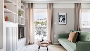 A modern living room with a green sofa, a wooden coffee table, and a TV on a white cabinet. There is a view of a balcony with outdoor furniture. - Iconic 1BR Apartment in Koukaki (Athens)