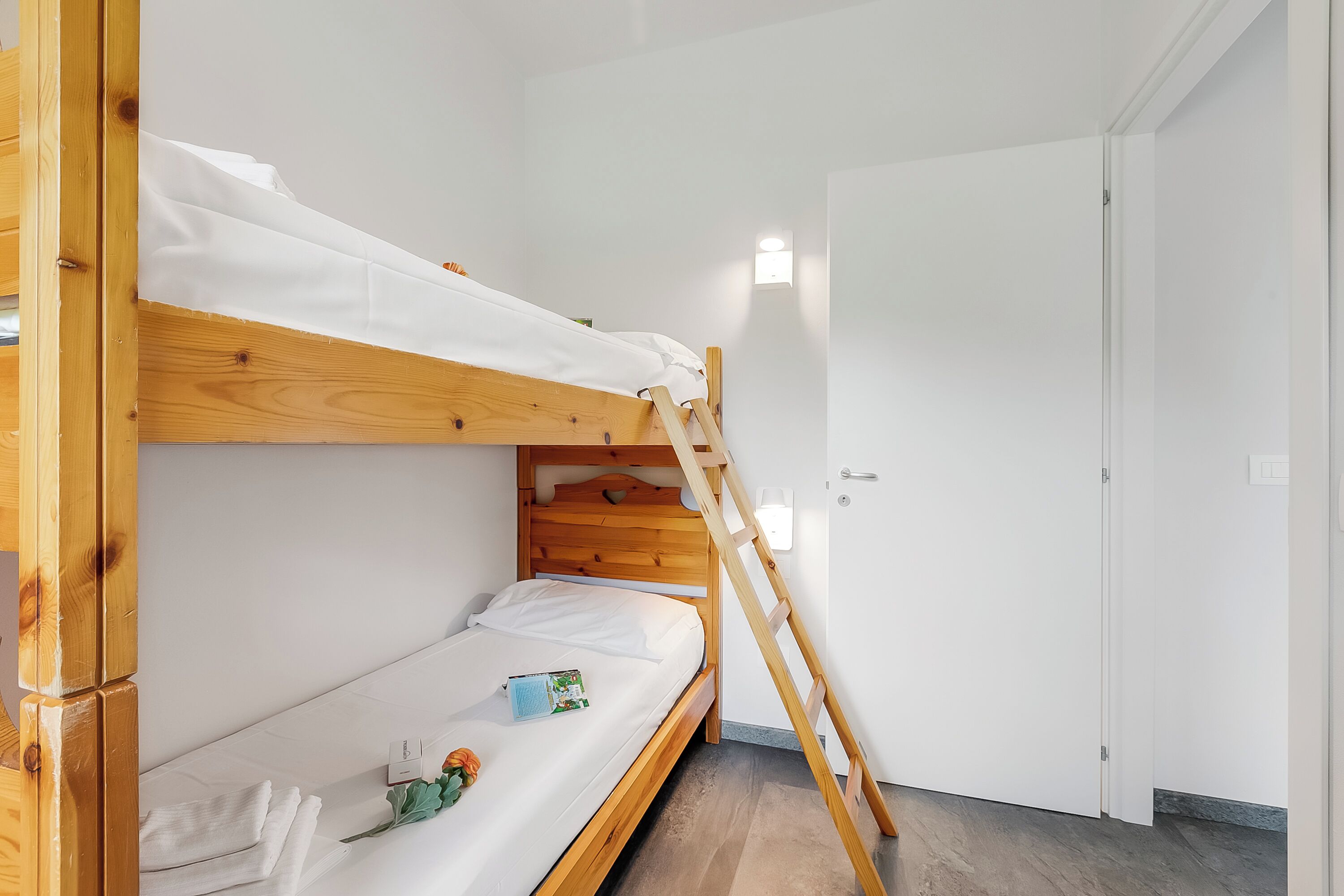 Ein Stockbett mit einer Holzleiter, weißer Bettwäsche und einem kleinen Nachttisch mit einem Buch und einer Blume.