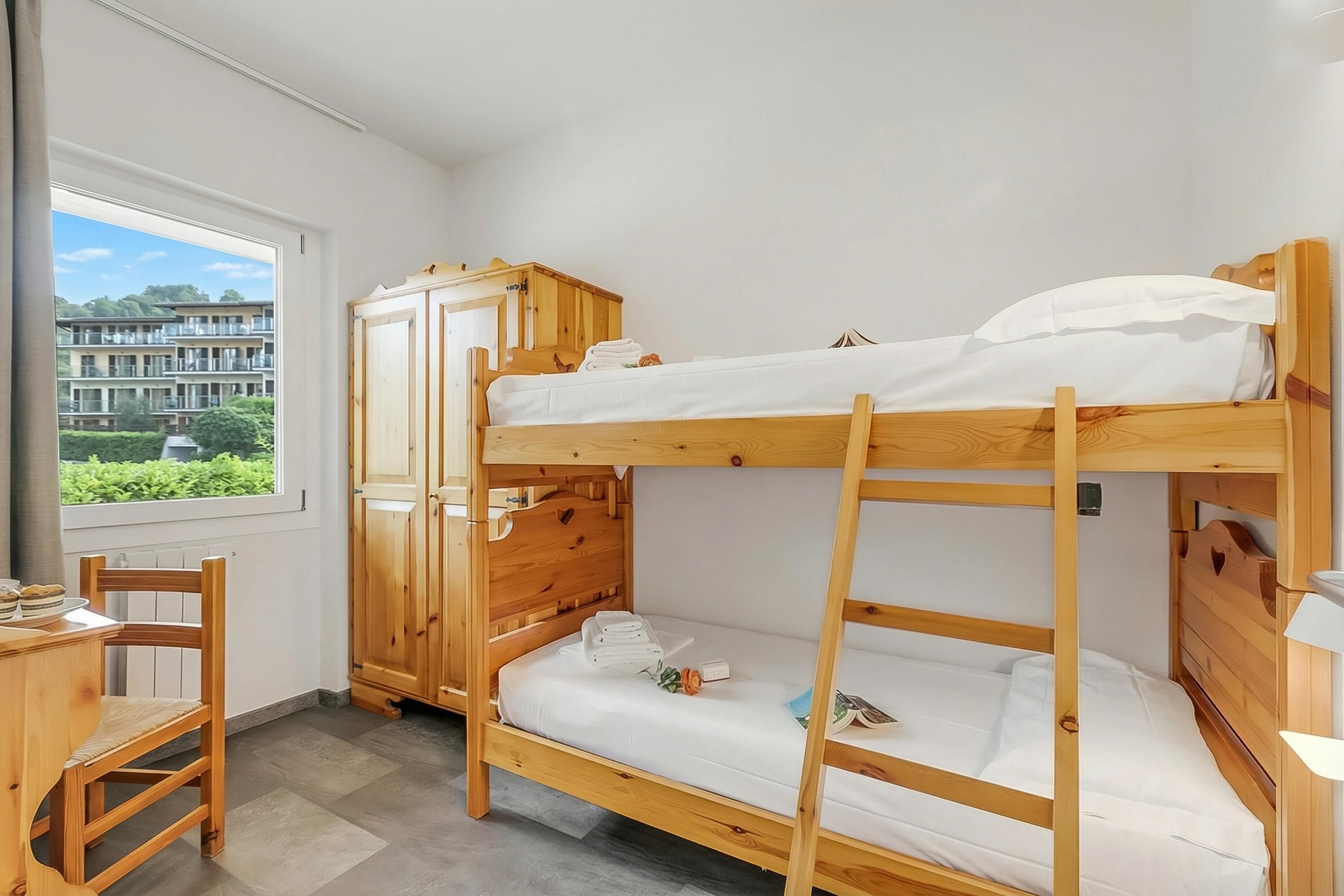 Ein Schlafraum mit Etagenbetten, einem Holzkopfteil, einem Schreibtisch mit Stuhl und Blick auf ein Gebäude durch ein Fenster.