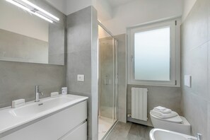 A modern bathroom with a large mirror, a white sink, a glass shower enclosure, and a toilet with a towel stack.