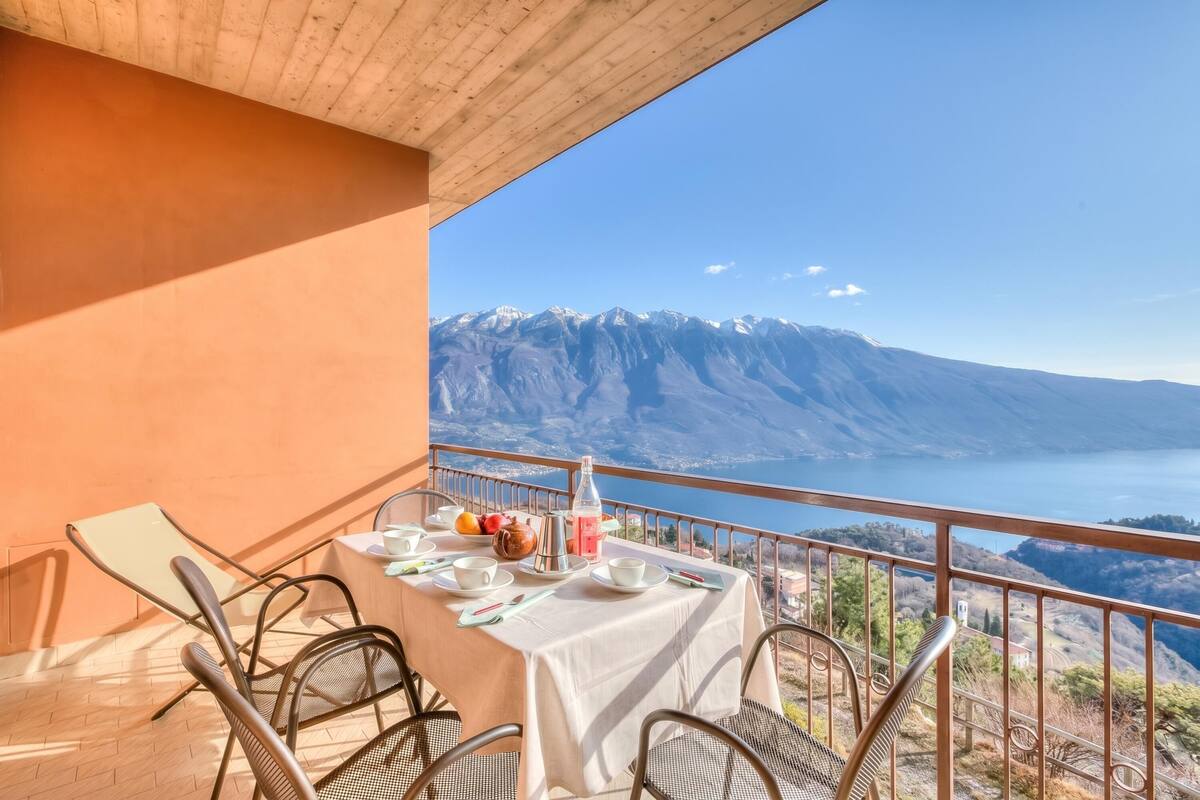 Mesa posta com xícaras, pratos e um jarro em uma varanda com vista para as montanhas.