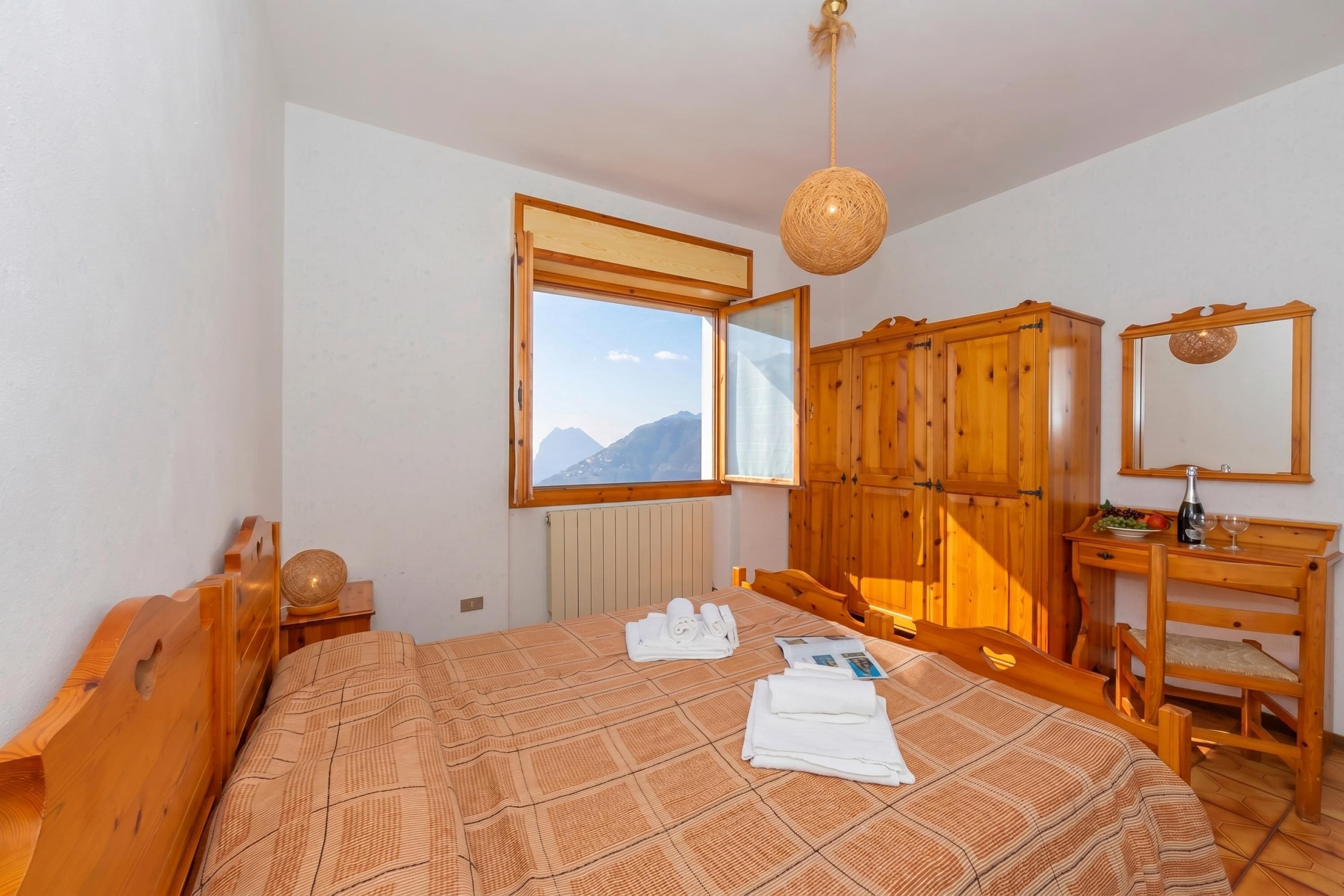 A bedroom with a bed, wooden wardrobe, a desk with a mirror, and a window with a mountain view.