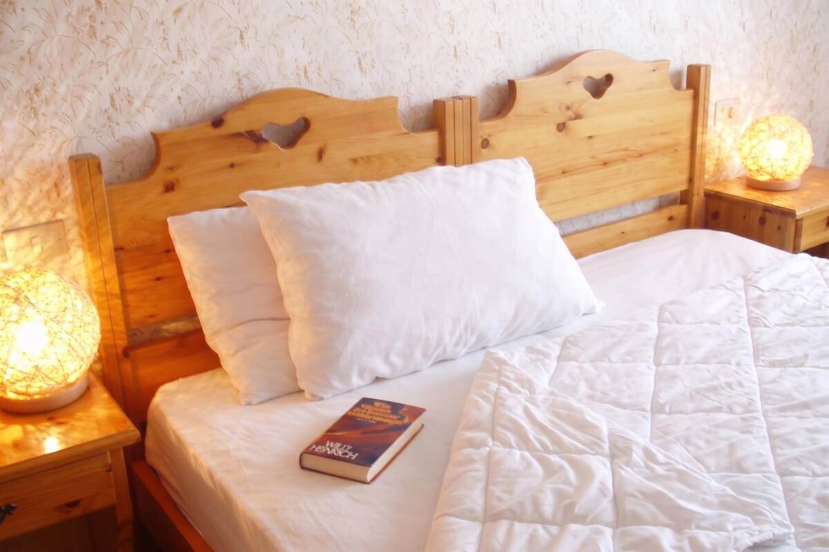 Uma cama de madeira com roupa de cama branca, um livro e duas mesas de cabeceira com abajures.