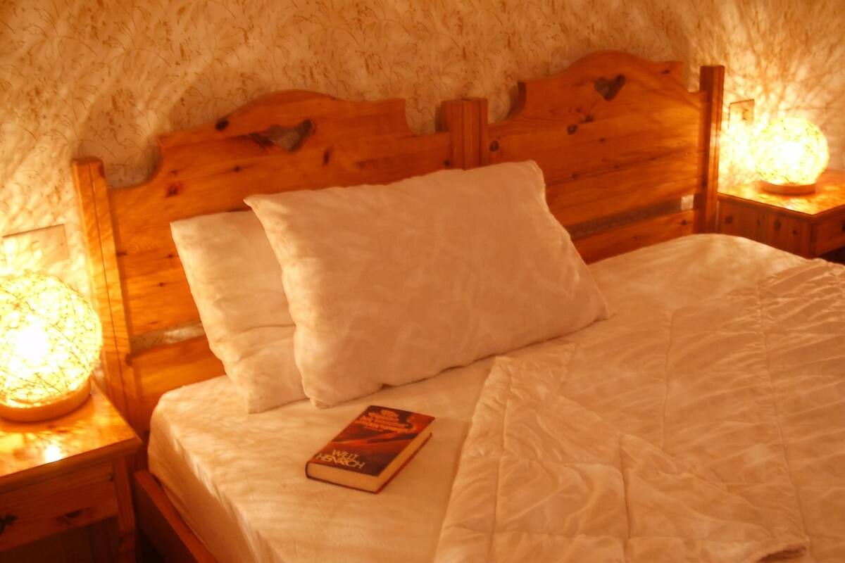 Uma cama de madeira com roupa de cama e almofadas brancas, um livro sobre a cama e duas luminárias de cabeceira.