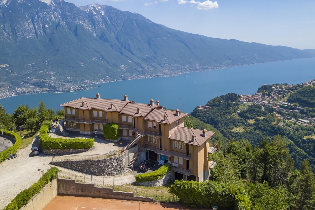 A large, multi-story house with a prominent balcony, situated on a hillside overlooking a lake and mountains.