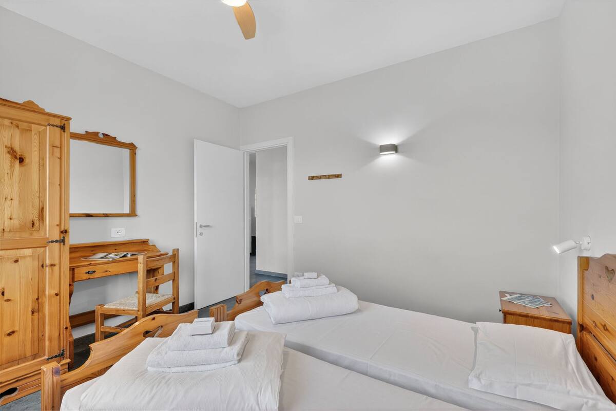 Quarto de hotel com duas camas, uma escrivaninha de madeira, um guarda-roupa e um ventilador de teto.