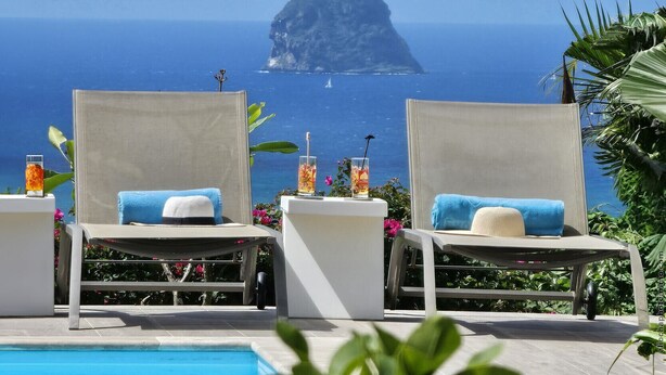 Zwei Liegestühle mit Handtüchern, ein Pool und Blick auf das Meer mit einer markanten Felsformation.