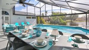 Un área de comedor al aire libre con vista a la piscina, un ventilador de techo y una mesa dispuesta con vasos y platos azules.