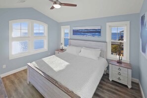 A bedroom with a large bed, two nightstands, a ceiling fan, and three windows offering views of the ocean. - Luxury Soundfront w/ Pool, Hot Tub, Elevator, Watersports Access, Media Room (Avon)