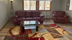 A living room with a brown leather sofa, a glass coffee table, and a patterned rug. There are two windows with blinds, a floor lamp, and a table lamp.