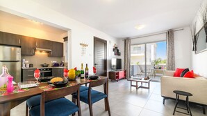 Una cocina moderna con armarios de madera oscura, un comedor con sillas tapizadas en azul y una sala de estar con sofá color beige y cojines rojos.