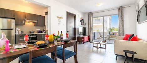 A modern kitchen with dark wooden cabinets, a dining area with blue upholstered chairs, and a living room with a beige sofa and red cushions.