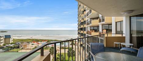 A balcony with a view of the beach and ocean, a pool, and outdoor seating.