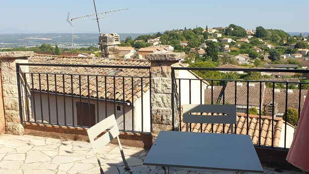 A terrace with stone flooring, a table, and a chair. There is a view of a town in the background. - Charming village house in the heart of the Luberon (Pierrevert)