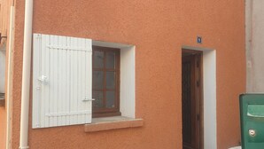 A building with a textured orange exterior, a white window with shutters, and a stone-paved entrance area. - Charming village house in the heart of the Luberon (Pierrevert)