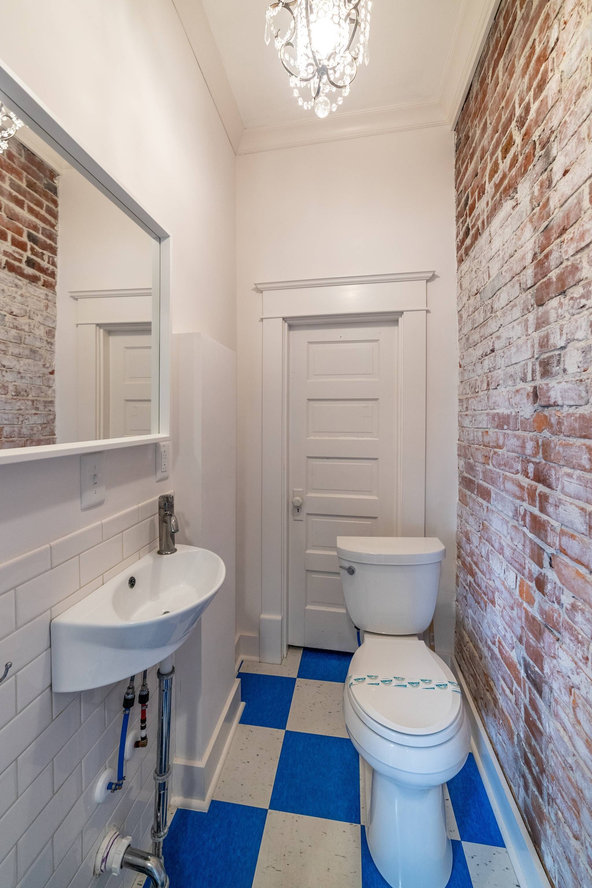 A bathroom with a white sink, a white toilet, a blue and white checkered floor, a brick wall, and a chandelier.