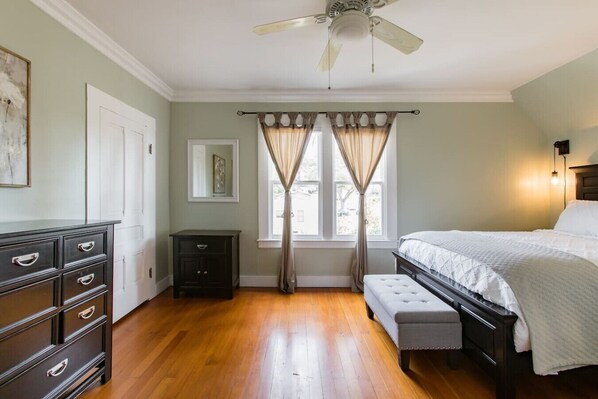 Un dormitorio con cama, mesita de noche, cómoda, ventilador de techo y ventana con cortinas.