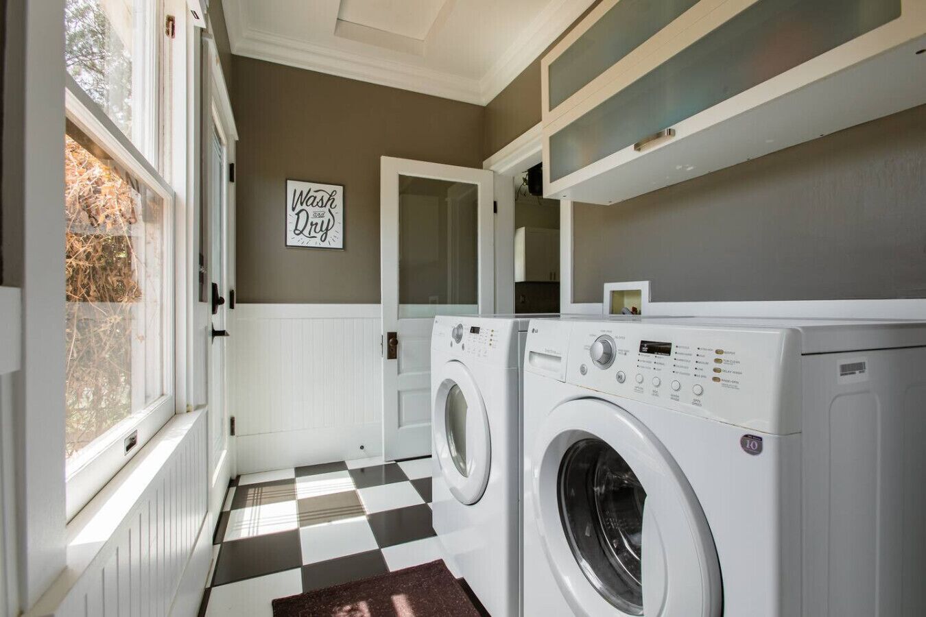 A laundry room with two white front-loading washers and dryers, a checkered floor, a 'Wash and Dry' sign, and a window with a view of trees.
